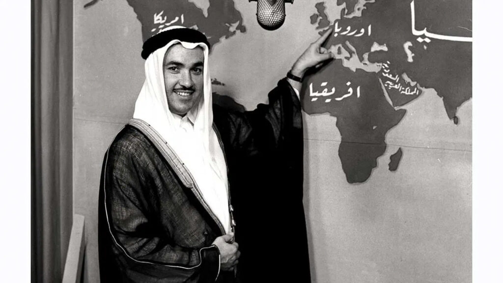 فهمي بصرامي يرتدي الزي السعودي والبشت ويشير إلى خريطة العالم خلفه يشرحها